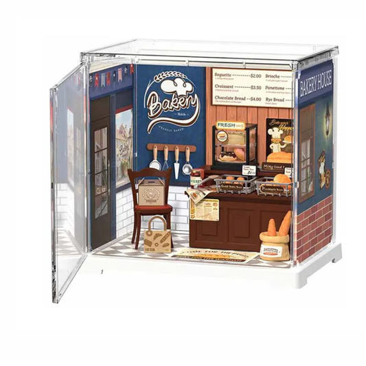 A miniature dollhouse model of a bakeshop storefront with an open side door, featuring tiny details like a counter, sign, chairs, and a basket on a white background.