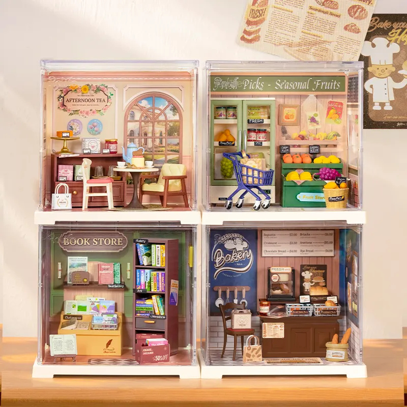 Four miniature dollhouse-style room sets displayed on a wooden surface against a cream wall. The sets include an afternoon tea room, a grocery store with a shopping cart, a book store, and a bakery.