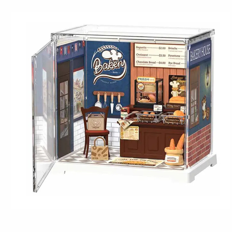 A miniature dollhouse model of a bakeshop storefront with an open side door, featuring tiny details like a counter, sign, chairs, and a basket on a white background.