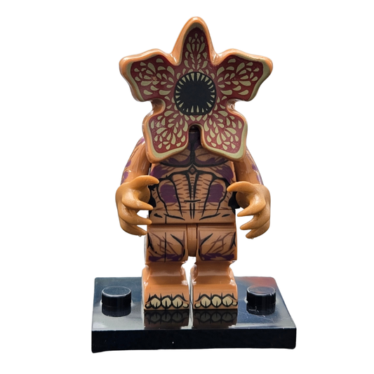 A toy minifigure of a flower-like monster with a large red and beige flower head, patterned brown torso, orange hands, and a black display base, shown on a white background.