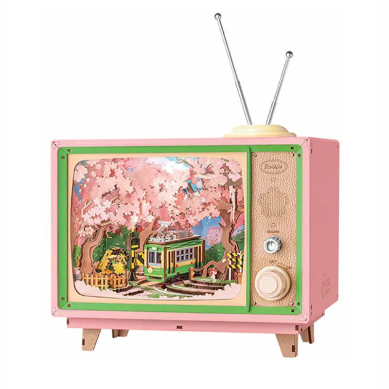 A pink vintage-style wooden TV toy with green trim, two metal antennas, and a colorful illustrated screen showing cherry blossoms and a green tram. The side panel has textured details, dials, and small wooden feet.