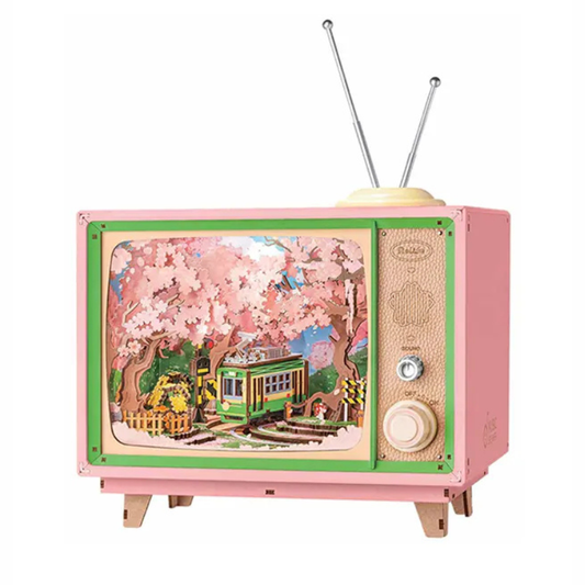 A pink vintage-style wooden TV toy with green trim, two metal antennas, and a colorful illustrated screen showing cherry blossoms and a green tram. The side panel has textured details, dials, and small wooden feet.