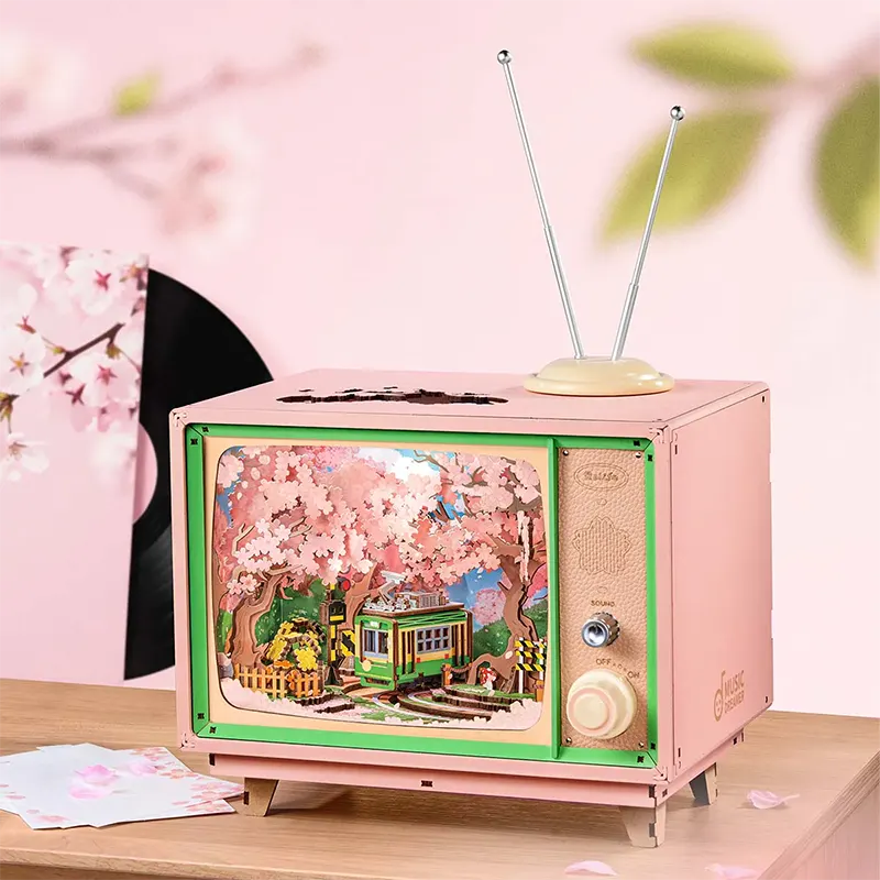 A pink vintage-style TV box model with green trim and two antenna rods, displayed on a wooden surface against a soft pink background with cherry blossom leaves and a vinyl record partially visible behind it.
