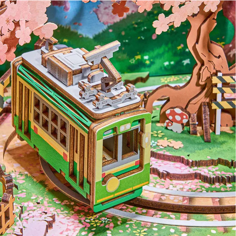 A 3D wooden model kit of a green and yellow electric tram on a track, set against a colorful forest backdrop with cherry blossoms, a tree, and mushrooms.