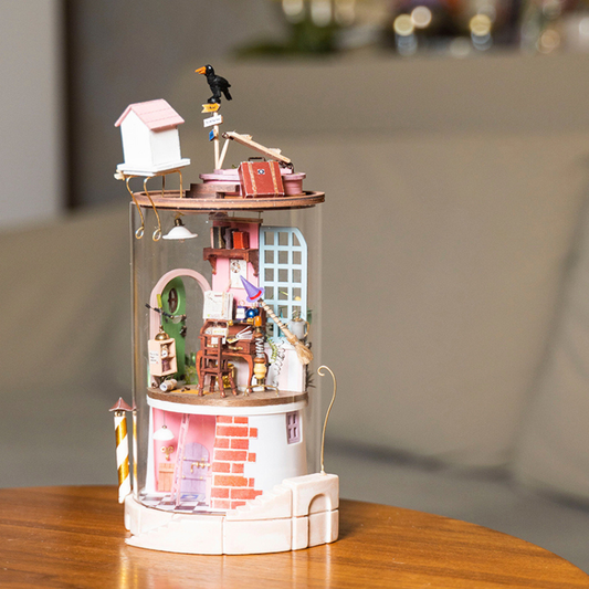 A miniature dollhouse-style decorative tower with a clear cylindrical body, pink and white base, tiny house, bird figurine, and detailed interior scenes, displayed on a wooden table with a softly blurred indoor background.