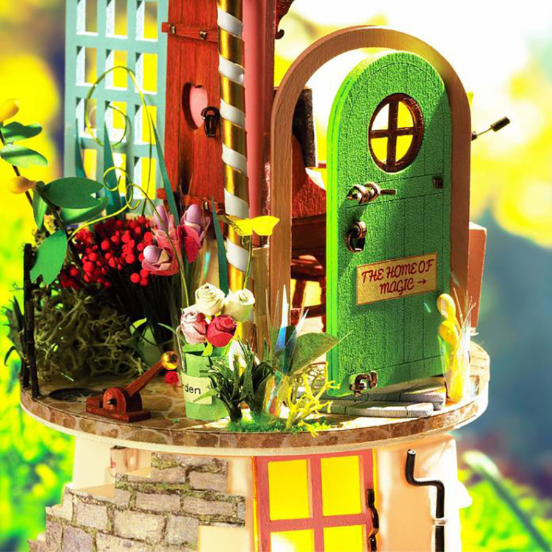 A miniature fairy house or dollhouse scene with a bright green door labeled 'THE HOME OF MAGIC' on a round table, surrounded by small flowers, plants, and whimsical architectural details against a blurred yellow-green background.